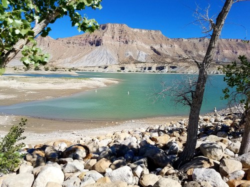 A serene landscape featuring a turquoise lake, rocky shore, and a backdrop of rugged mountains under a clear blue sky.