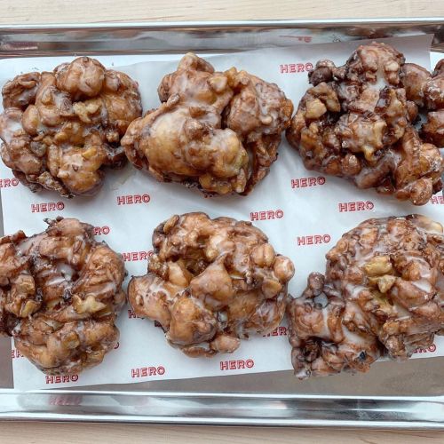 A tray of six glazed apple fritters with a crispy texture and chunks of apple, resting on branded paper.