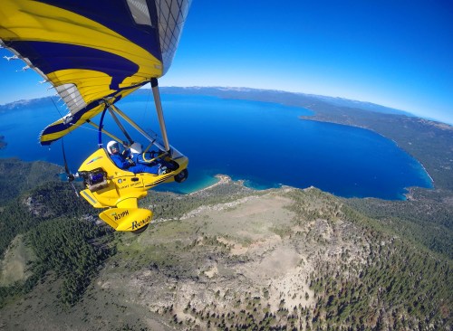 A yellow ultralight aircraft flying over a scenic lake surrounded by mountains and forests under a clear blue sky.