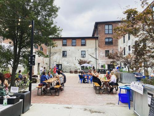 A lively outdoor space with people dining at tables, surrounded by brick buildings and greenery under a cloudy sky.