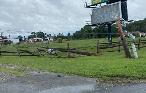 A damaged fence and downed power lines in a grassy area, with a billboard in the background and cloudy skies above.