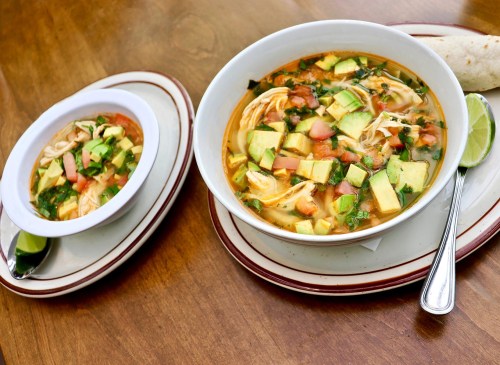 Two bowls of chicken soup with avocado, tomatoes, and cilantro, served with lime wedges on a wooden table.