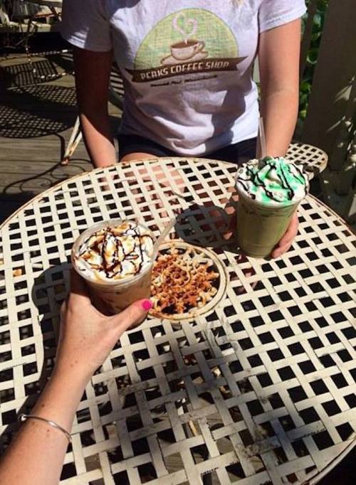 Two iced coffee drinks with whipped cream and a bowl of snacks on a table, with a person holding one drink.