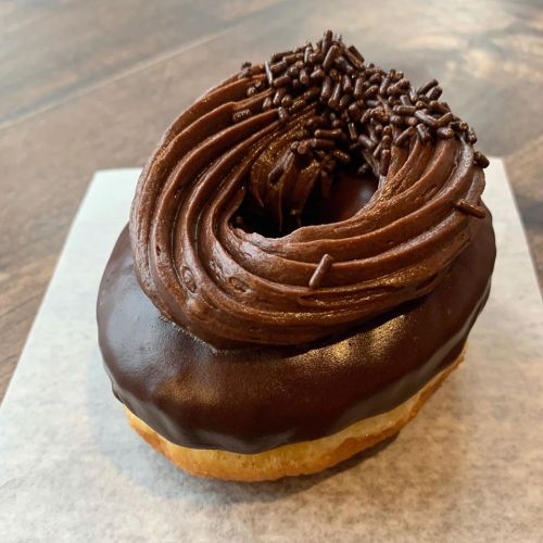 A chocolate frosted donut topped with chocolate frosting and sprinkles, resting on a piece of parchment paper.