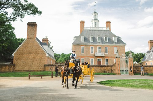 A horse-drawn carriage approaches a historic brick building with a tall steeple, surrounded by greenery.