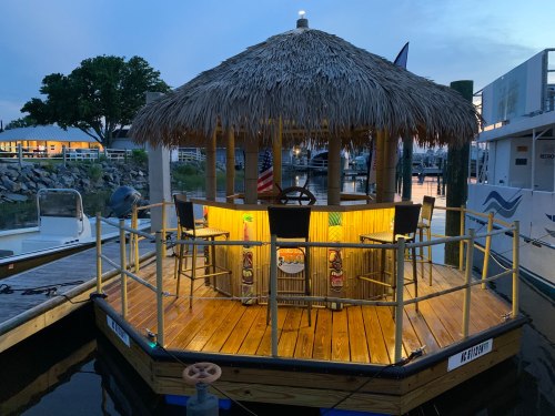 A tiki bar on a dock, featuring a thatched roof, illuminated seating, and a view of the water at dusk.