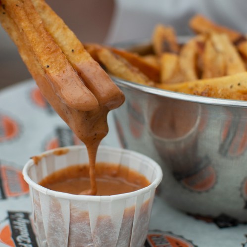 A hand dips a crispy fry into a small cup of thick, savory sauce, with a bowl of fries in the background.