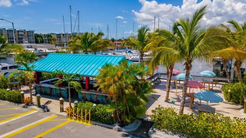 A vibrant waterfront scene featuring a blue-roofed bar, palm trees, and colorful umbrellas by a marina with sailboats.