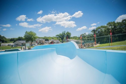 A blue water slide at a park, with picnic tables and trees in the background under a clear blue sky.