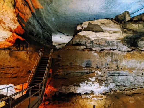 A dimly lit cave interior with a staircase leading up to a rocky ceiling, showcasing natural formations and colorful lighting.