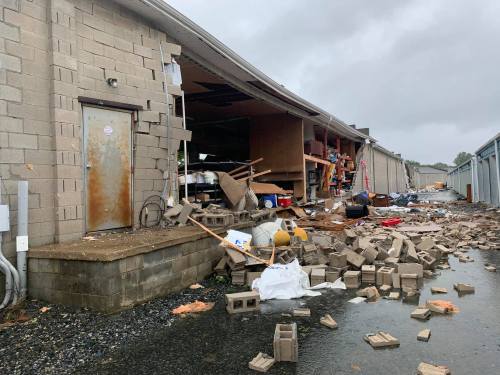 A damaged building with debris scattered around, including cinder blocks and scattered items, under a cloudy sky.