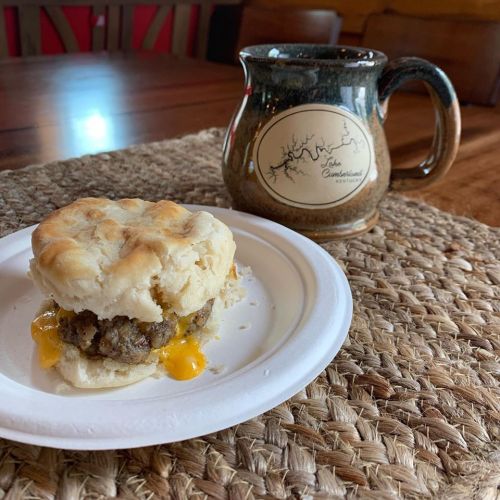 A biscuit sandwich with sausage and cheese on a paper plate, next to a coffee mug on a woven placemat.