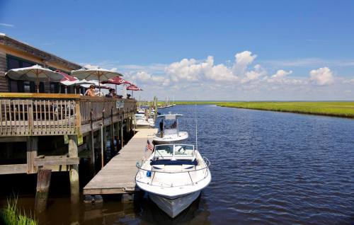 A boat docked by a wooden pier next to a restaurant, with lush green marshland and blue skies in the background.