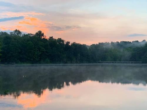 A serene lake at dawn, reflecting colorful clouds and surrounded by lush trees, with mist rising from the water.
