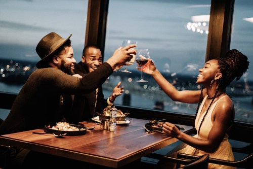 Three friends toast with glasses of wine at a stylish restaurant, enjoying a scenic view and a lively atmosphere.