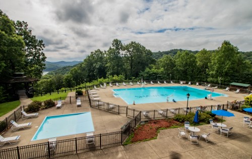 A scenic outdoor pool area surrounded by trees, featuring lounge chairs and a view of distant mountains under a cloudy sky.