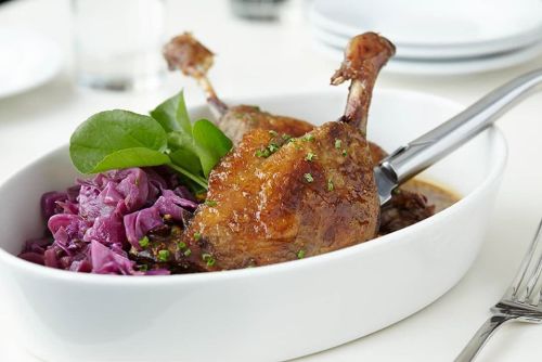 A dish featuring two crispy duck legs served with purple cabbage and garnished with fresh greens.
