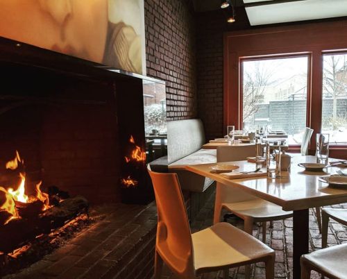 Cozy restaurant interior with a fireplace, wooden tables, and large windows overlooking a snowy landscape.