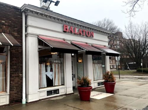 Exterior of Balaton restaurant with red signage, awnings, and potted plants on a rainy day.