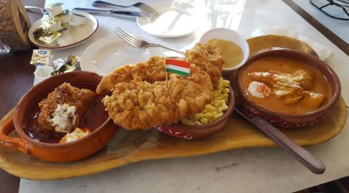 A wooden platter with fried chicken, a meat dish in sauce, and pasta, garnished with a small Hungarian flag.