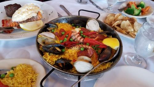 A vibrant seafood paella with mussels, clams, shrimp, red peppers, peas, and lemon, served with sides of vegetables and potatoes.