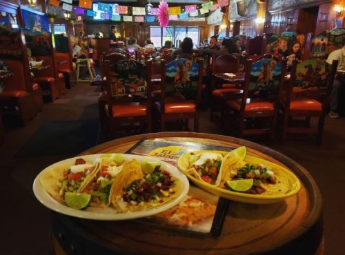 Three colorful tacos with toppings on a plate, set on a wooden table in a lively restaurant filled with diners.