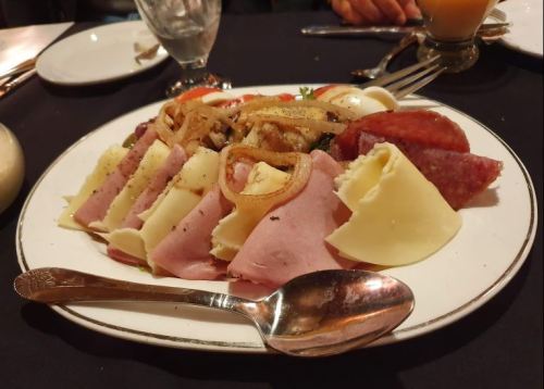 A platter of assorted meats and cheeses, garnished with onions, served on a white plate with a silver spoon.