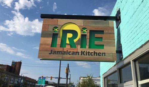 Sign for "iRiE Jamaican Kitchen" featuring a colorful design with Jamaican flag elements against a blue sky.