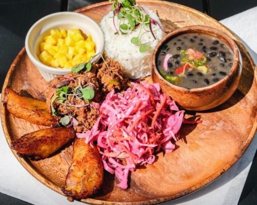 A wooden plate with rice, black bean soup, pulled meat, pickled cabbage, fried plantains, and mango chunks.