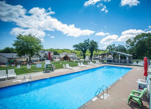 A clear blue swimming pool surrounded by lounge chairs, with green trees and buildings in the background under a bright sky.