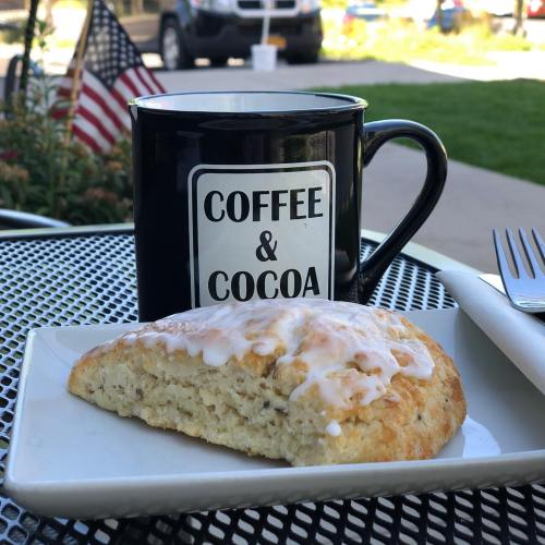 A black mug labeled "Coffee & Cocoa" beside a glazed scone on a white plate, set on a table outdoors.