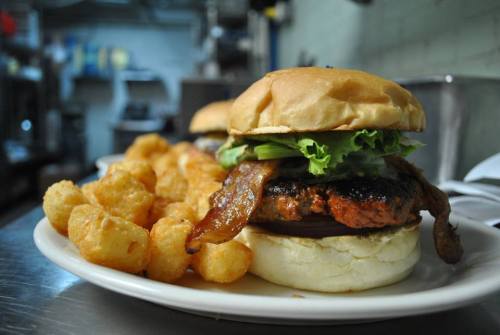 A close-up of a burger with lettuce and bacon, served with a side of crispy tater tots on a plate.