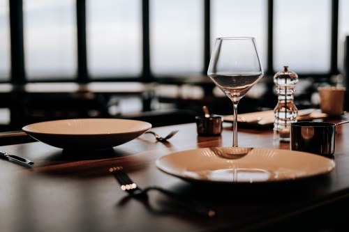 A dining table set with two plates, a wine glass, a pepper mill, and a small container, with large windows in the background.