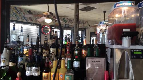 A bar counter filled with various bottles of liquor, a slushy machine, and a mirror reflecting the bar's interior.