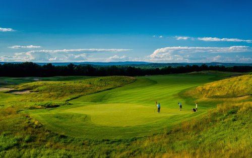 A scenic golf course with players on the green, surrounded by lush grass and a clear blue sky.