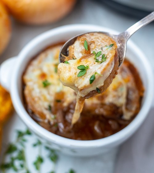 A spoon lifting a cheesy, herb-topped serving of French onion soup from a white bowl.