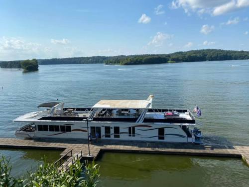 A houseboat docked on a calm lake, surrounded by greenery and blue skies with fluffy clouds.