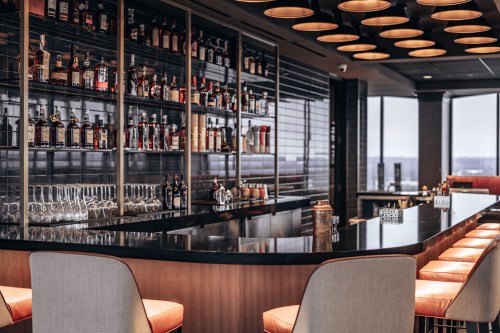 A stylish bar with a sleek black countertop, shelves of liquor bottles, and modern seating in a well-lit space.