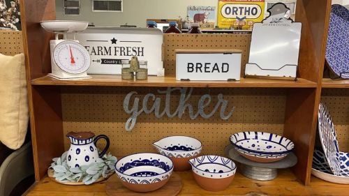 A wooden shelf displays decorative items, including bowls, a scale, and signs that say "gather" and "bread."