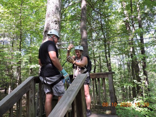 Two people in helmets prepare for a zip line in a wooded area, standing on a wooden platform.