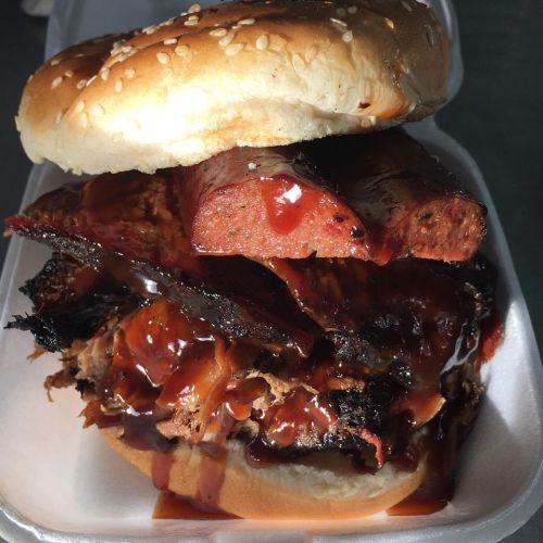 A stacked sandwich featuring layers of smoked meats, including brisket and sausage, topped with barbecue sauce on a sesame bun.