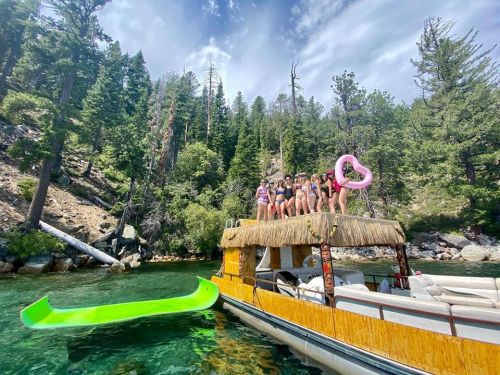 A group of people in swimsuits poses on a boat surrounded by trees and clear water, with a green float nearby.