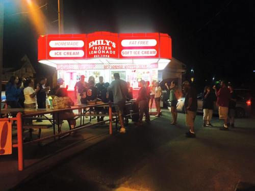 A crowd gathers at Emily's Frozen Lemonade stand at night, enjoying ice cream and lemonade under bright red signage.