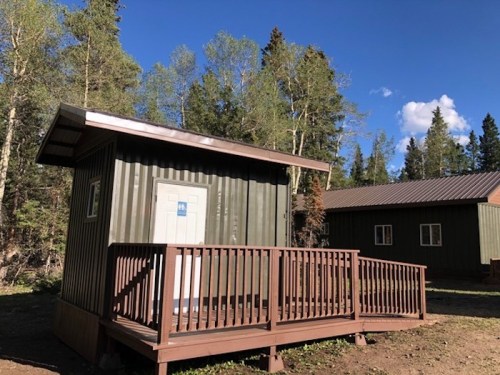 A small green cabin with a wooden deck, surrounded by trees and blue sky.