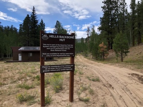 Sign for Hell's Backbone Hut, part of the Aquarius Trail Hut System, surrounded by trees and a dirt path.