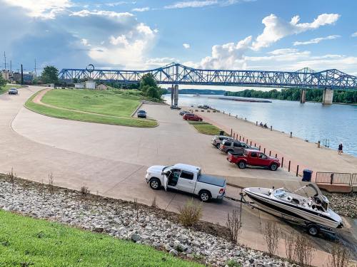 A truck with a boat is parked by a river, with a bridge in the background and a grassy area nearby.