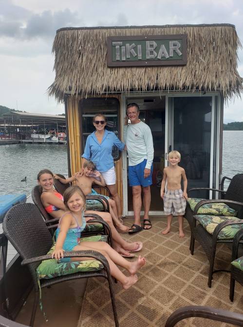 A family poses outside a tiki bar on a dock, with children sitting and smiling, enjoying a sunny day by the water.
