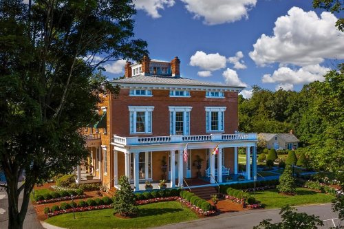 A grand brick house with white columns, surrounded by lush greenery and colorful flower beds under a blue sky with clouds.