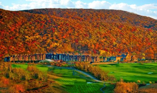 A scenic view of a resort nestled at the base of a colorful autumn mountain, with green fields in the foreground.