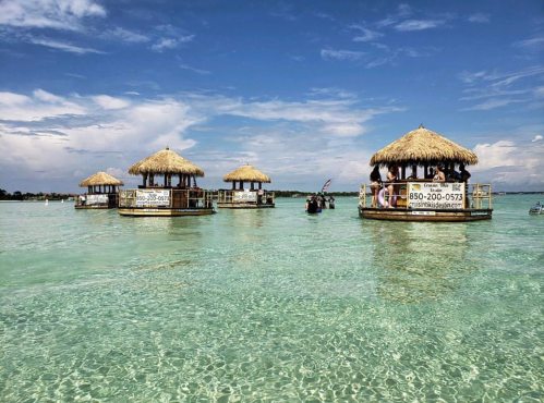 Three thatched-roof huts on stilts in clear turquoise water under a blue sky with scattered clouds.